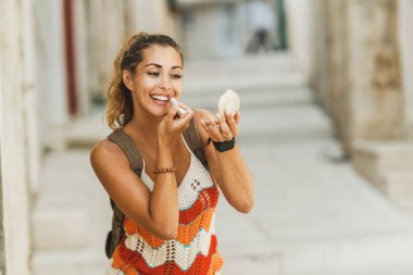 A young smiling woman using hand mirror and applying lipstick while enjoying a summer vacation.