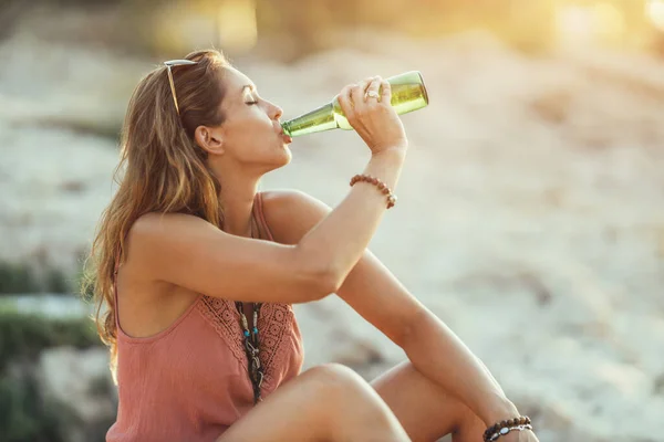 An attractive young woman drinking beer while enjoying a summer vacation on the beach by the sea.
