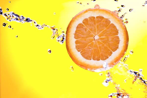 View of piece of orange getting splashed and glass of juice on back — Stock Photo, Image