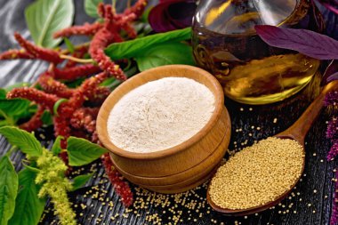 Bir kasede Amaranth unu, bir kaşıkta tohumlar ve sürahide yağ, yapraklar ve kahverengi, yeşil, mor çiçekler koyu ahşap arka planda.