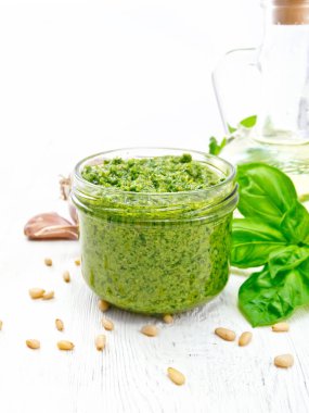 Cam bir kavanozda pesto sosu, fesleğen, çam fıstığı, sarımsak ve zeytinyağı beyaz ahşap arka planda bir sürahi içinde