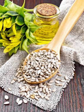 Çuval bezinde ve masada kaşık kaşık içinde ayçiçeği tohumları, cam kavanozda yağ ve ahşap arka planda ayçiçeği çiçeği.
