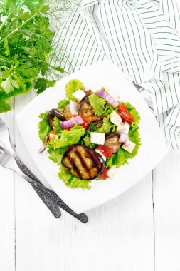 Kızarmış patlıcan salatası, taze soğanlı domates ve tuzlu feta peyniri, sebze yağı ve soya sosuyla tatlandırılmış, tabakta marul, havlu, çatal ve maydanoz, ahşap tahta arka planda.