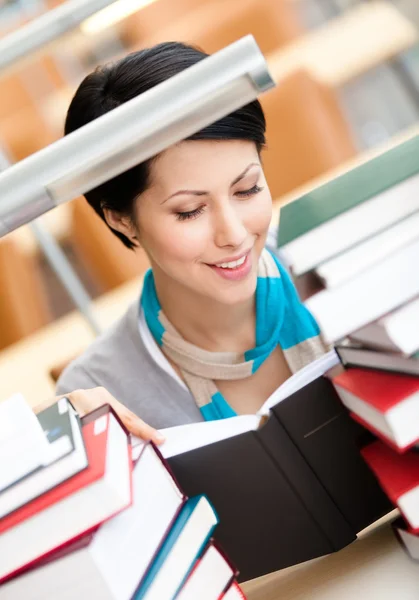 Reading book female student surrounded with piles of books sitting at the desk at the library — Stock Photo, Image