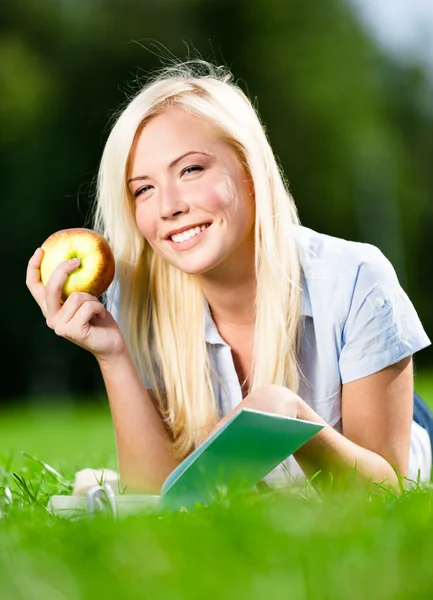 Mulher com maçã lê livro deitado na grama verde — Fotografia de Stock