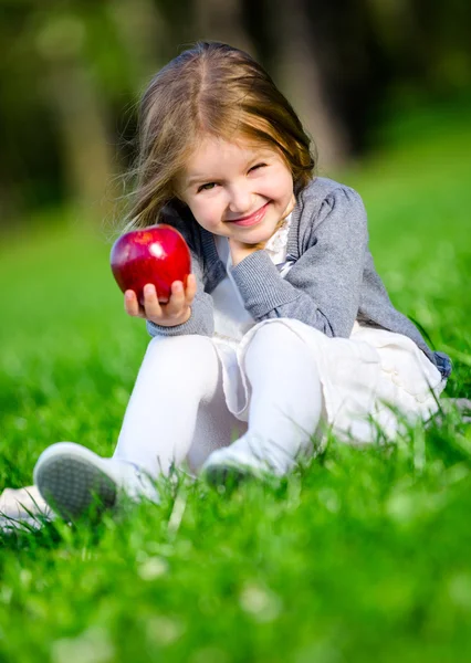 Yeşil çimenlerin üzerinde oturan kırmızı elma ile küçük kız — Stok fotoğraf