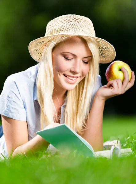 Chica en sombrero de paja con manzana lee libro sobre la hierba verde — Foto de Stock