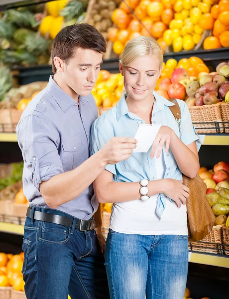 Счастливая пара со списком покупок на фоне стопок фруктов — стоковое фото
