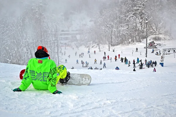 Sportsman en suite verte avec skateboard assis sur neige blanche . — Photo