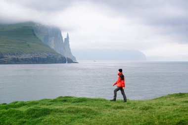 Vagar, Faroe Adaları 'ndaki Vagar köyü yakınlarındaki Cadıların Parmağı veya Trollkonfingur' un dağlarına bakan kırmızı ceketli bir turist.