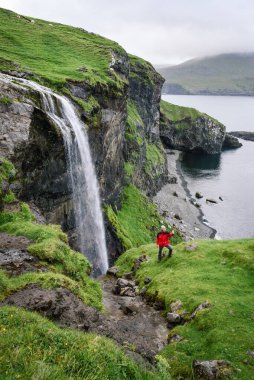 Kırmızı ceketli bir gezgin, Faroe Adaları 'ndaki Vagar Adası' ndaki Skardsafossur şelalesinin yanında telefonla selfie çekiyor.