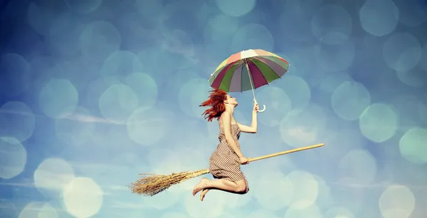 Молодая рыжая ведьма на метле летит в небе с зонтиком — стоковое фото