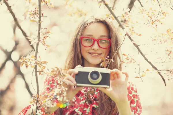 Menina adolescente em óculos com câmera vintage perto da árvore da flor — Fotografia de Stock