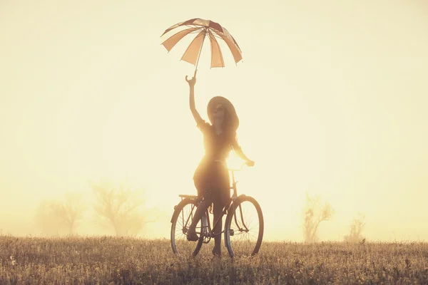 Menina com guarda-chuva em uma bicicleta no campo ao nascer do sol — Fotografia de Stock