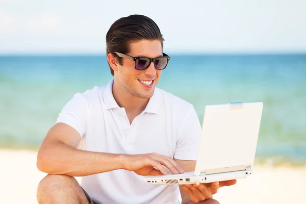 Jovem bonito com laptop no fundo da praia — Fotografia de Stock