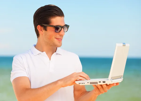 Beau jeune homme avec ordinateur portable à fond de plage — Photo