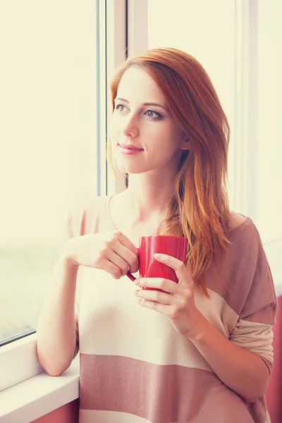 Beautiful young woman with cup over window. — Stock Photo, Image