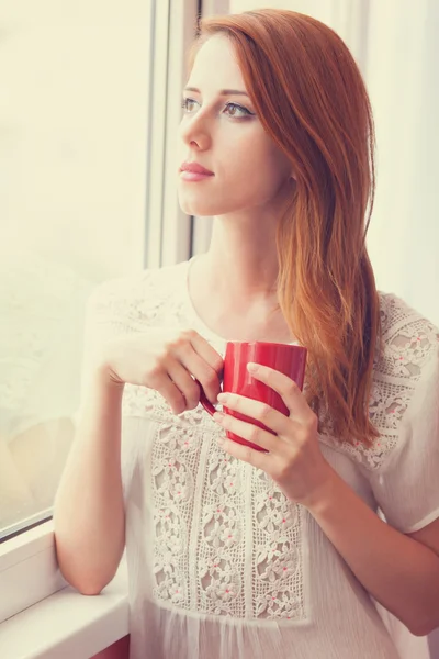 Beautiful young woman with cup over window. — Stock Photo, Image