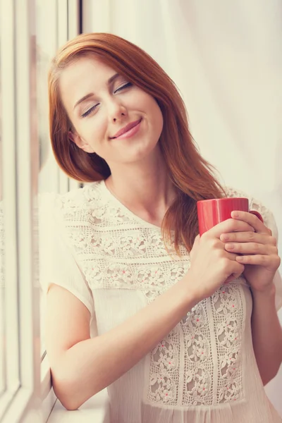 Beautiful young woman with cup over window. — Stock Photo, Image