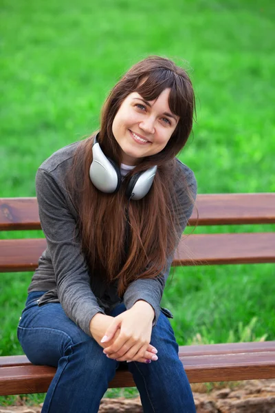 Bruna ragazza con le cuffie seduto alla panchina nel parco . — Foto Stock