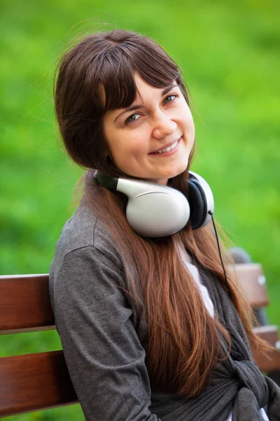 Bruna ragazza con le cuffie seduto alla panchina nel parco . — Foto Stock