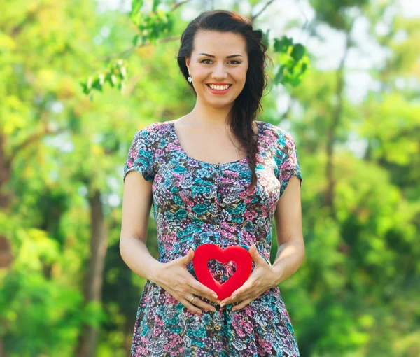 Jovem mulher grávida bonita com coração de brinquedo no parque — Fotografia de Stock