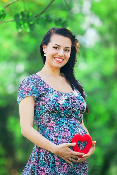 Young beautiful pregnant woman with toy heart in the park — Stock Photo, Image