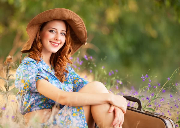 Рыжая девушка с чемоданом сидит на осенней траве — стоковое фото