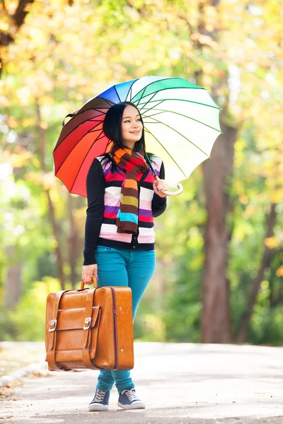 Chica morena sosteniendo maleta y paraguas en el callejón de otoño en t — Foto de Stock