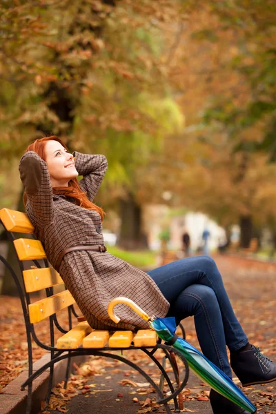Στυλ κοκκινομάλλα κοπέλα καθόταν στο παγκάκι στο πάρκο φθινόπωρο. — Φωτογραφία Αρχείου