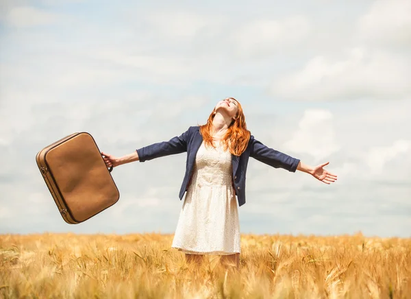 Redhead girl with suitcase at spring wheat field. — Stock Photo, Image