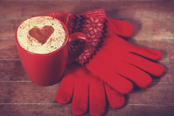 Gloves and cup with coffee and shape of the cacao heart on it. — Stock Photo, Image