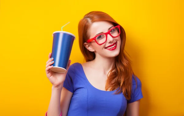 Rödhårig tjej med drink och glasögon på gul bakgrund. — Stockfoto