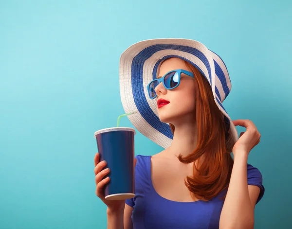 Κοκκινομάλλα κοπέλα με ποτό και γυαλιά ηλίου σε μπλε φόντο — Φωτογραφία Αρχείου