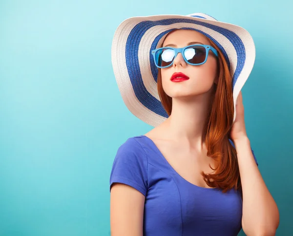 Roodharige meisje met zonnebril en hoed op blauwe achtergrond. — Stockfoto