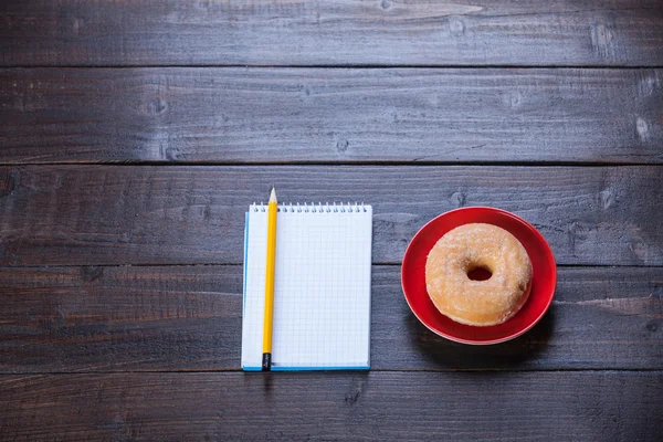 Cuaderno, rosquilla y lápiz sobre mesa de madera . —  Fotos de Stock