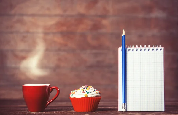 Cup of coffee with cupcake and notebook on wooden background. — Stock Photo, Image