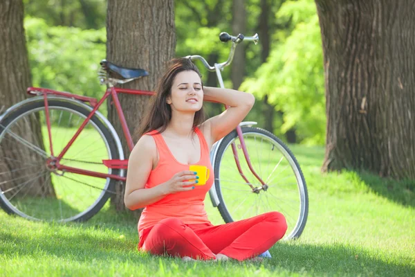 Brunette girl with cup of coffee in the park and with bicycle on — Stock Photo, Image