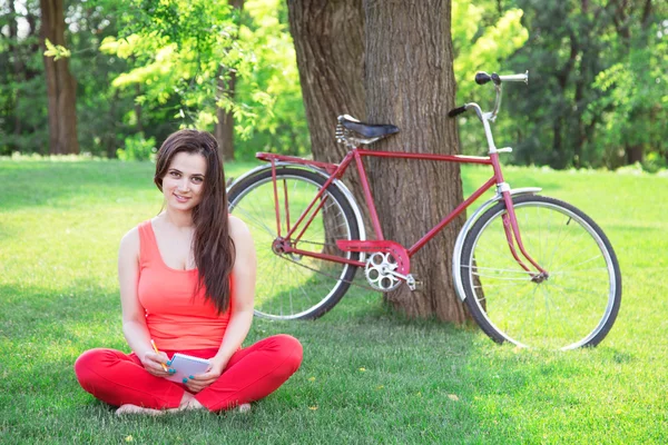 Brunette girl with notebook in the park and with bicycle on back — Stock Photo, Image