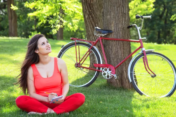 Brunette girl with notebook in the park and with bicycle on back — Stock Photo, Image