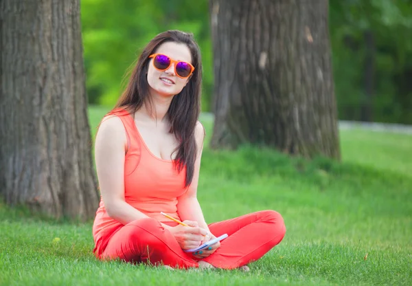 Chica morena con cuaderno sobre hierba verde en el parque . — Foto de Stock