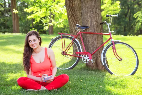 Brunette girl with notebook in the park and with bicycle on back — Stock Photo, Image