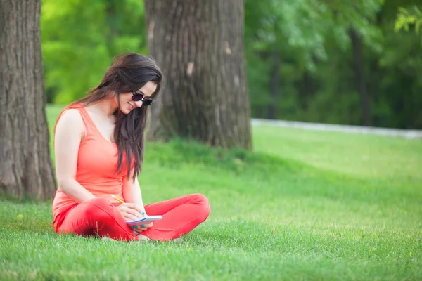 Brunettee flicka med anteckningsboken på grönt gräs i parken. — Stockfoto