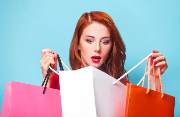 Surprised redhead girl with shopping bags on blue background. — Stock Photo, Image