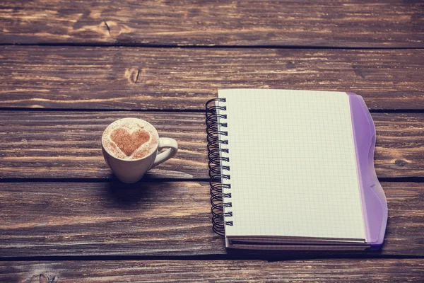 Taza de café con forma de corazón y cuaderno en mesa de madera . — Foto de Stock