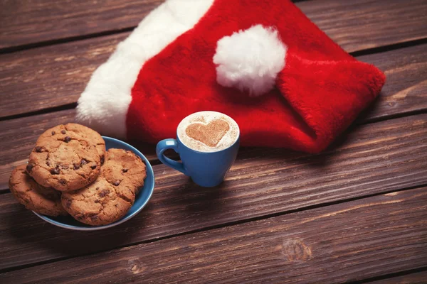 Cookie i filiżankę kawy z kapelusz Świętego Mikołaja na drewnianym stole. — Zdjęcie stockowe