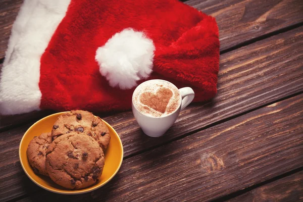 Plätzchen und Tasse Kaffee mit Weihnachtsmütze auf Holztisch. — Stockfoto