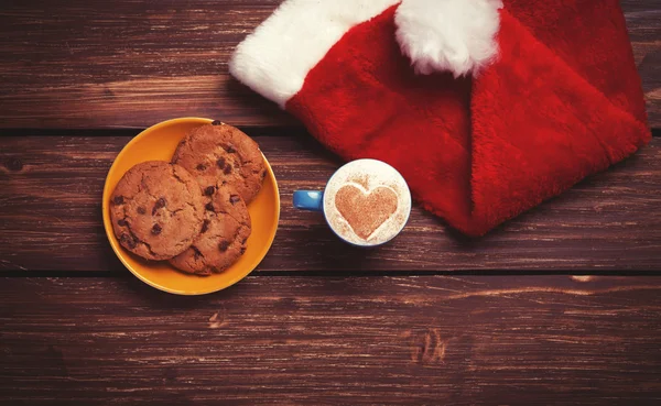 Plätzchen und Tasse Kaffee mit Weihnachtsmütze auf Holztisch. — Stockfoto