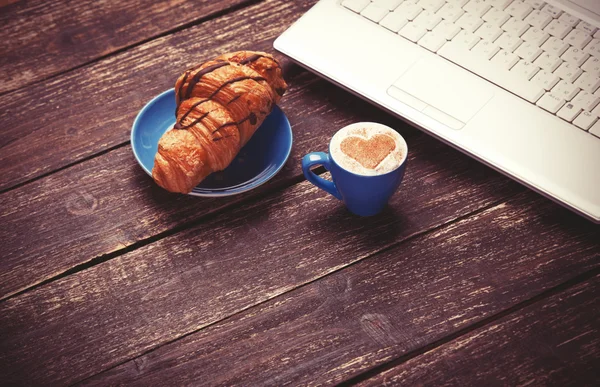 Croissant och en kopp kaffe med laptop på träbord. — Stockfoto