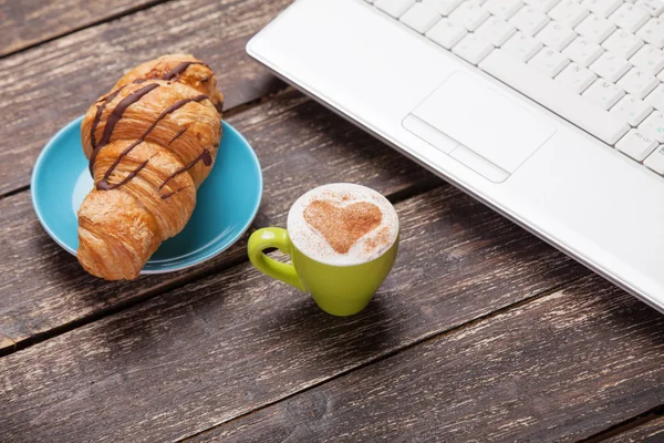 Croissant and cup of coffee with laptop on wooden table. — Stock Photo, Image